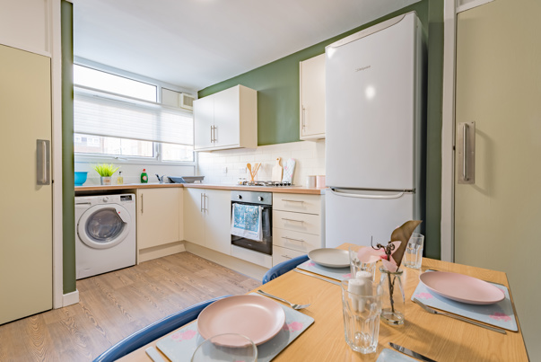 Image of a student house kitchen with a washing machine, fridge, oven, cream cabinets and green doors and a dining table which is set.